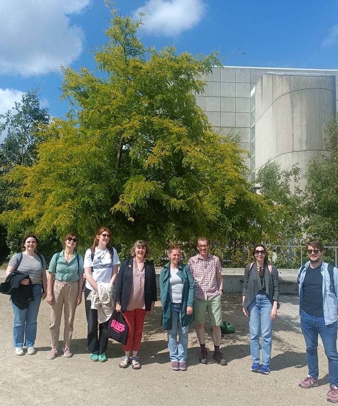 Earth Institute Writing Retreat 2024 group shot showing a group of people outdoors next to a vividly green leafed tree and building in the sunshine